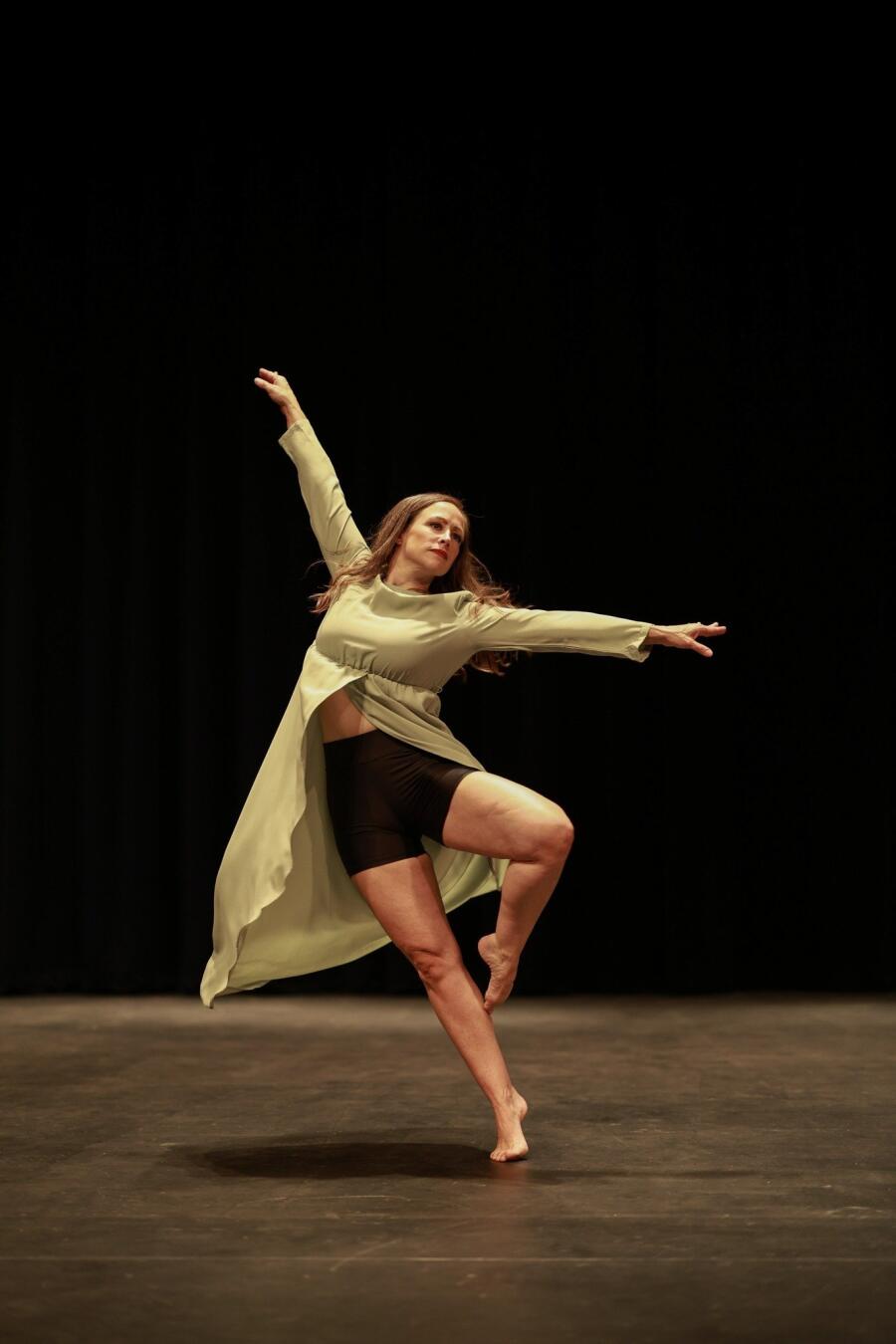 A dancer on a bare stage extending one arm and one leg into an arabesque under a single warm light.