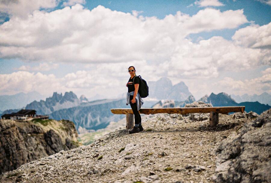 A hiker standing beside a wooden bench on a rocky ridge, with jagged peaks and cloud behind.