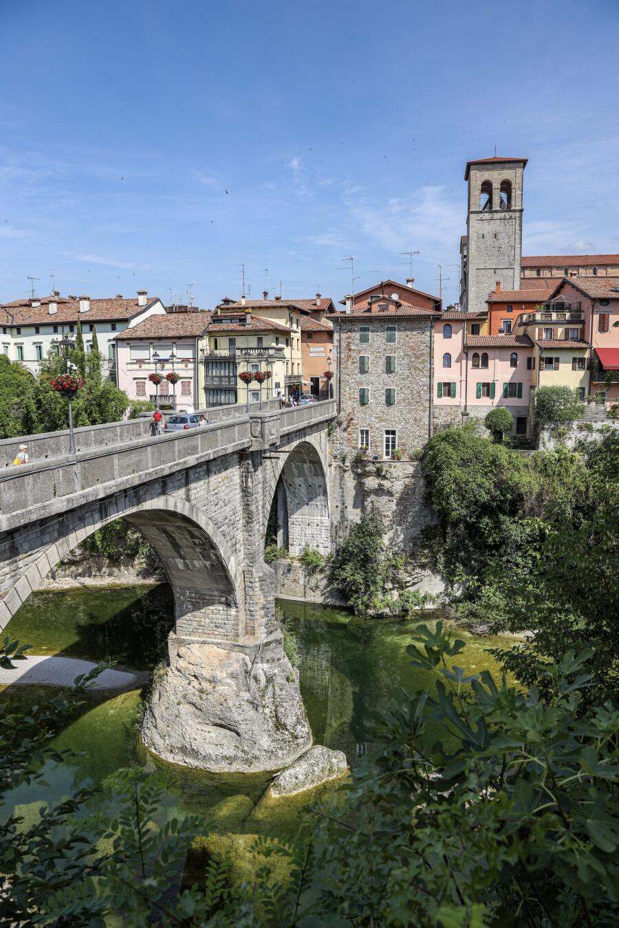 A stone arch bridge spanning a green river gorge below a town of tall houses and a bell tower.