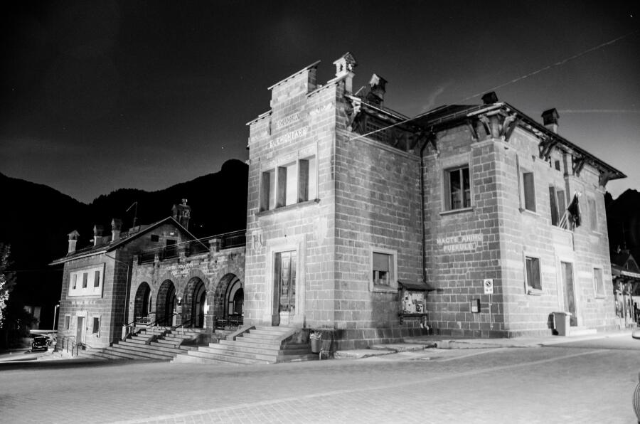 Black and white photograph of a stone civic building with an arcaded ground floor on a corner.