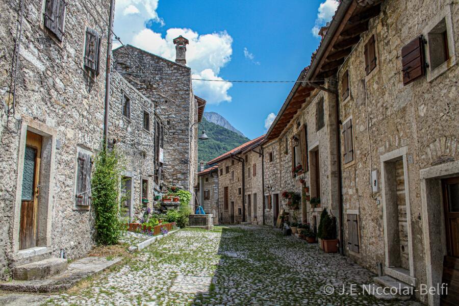 A narrow cobbled street between stone houses with shutters and window boxes, opening to a mountain beyond.