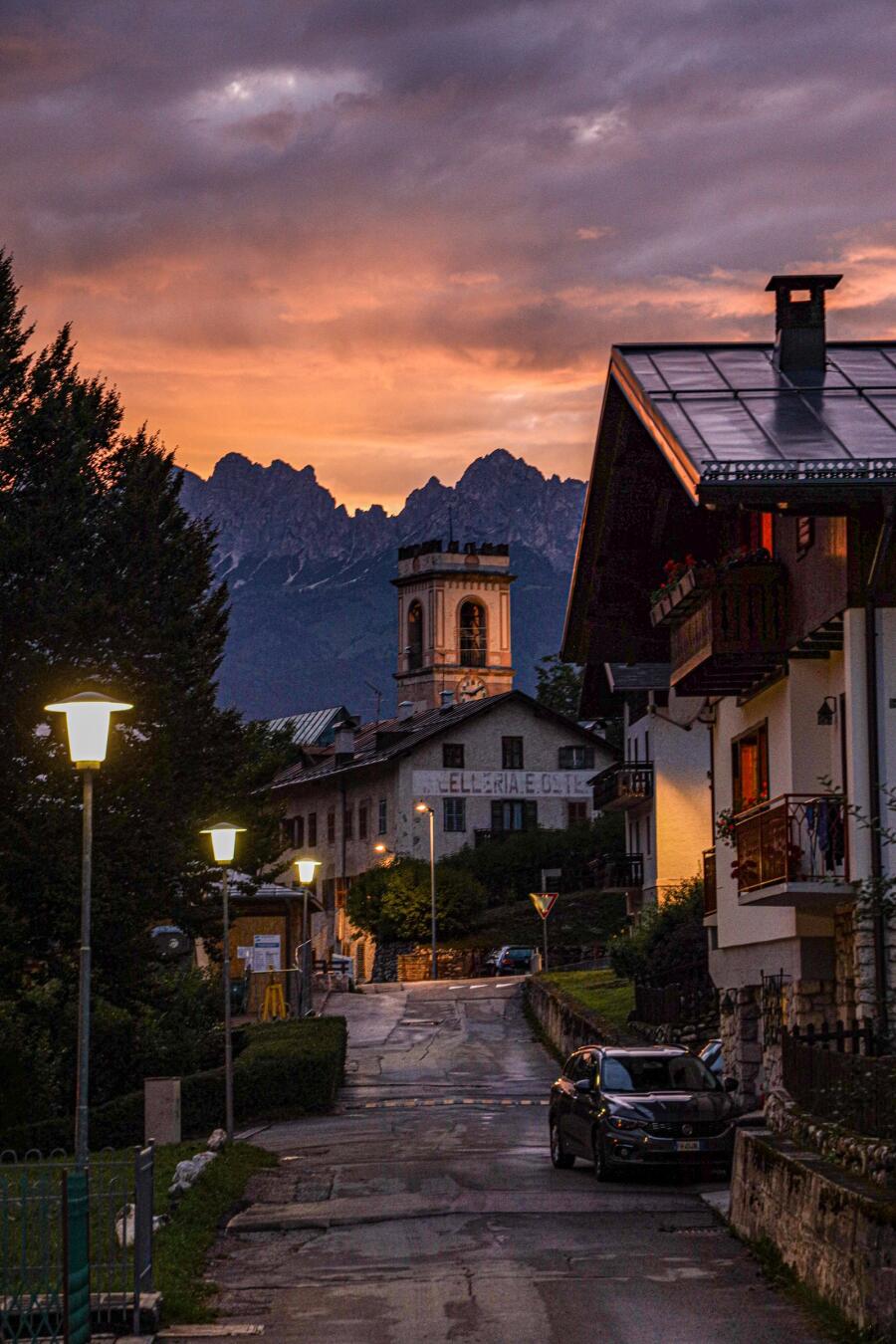 A village street at dusk with lit windows and a bell tower, under an orange and grey sky.