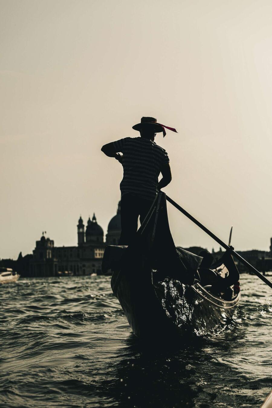 A gondolier silhouetted against pale water and a distant skyline, leaning into the oar.