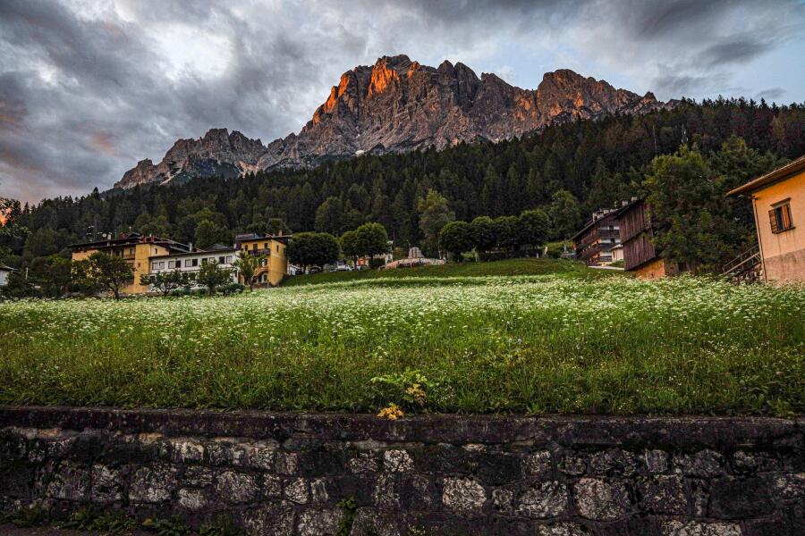 Warm evening light striking a rock ridge above a village, with a mown meadow in the foreground.
