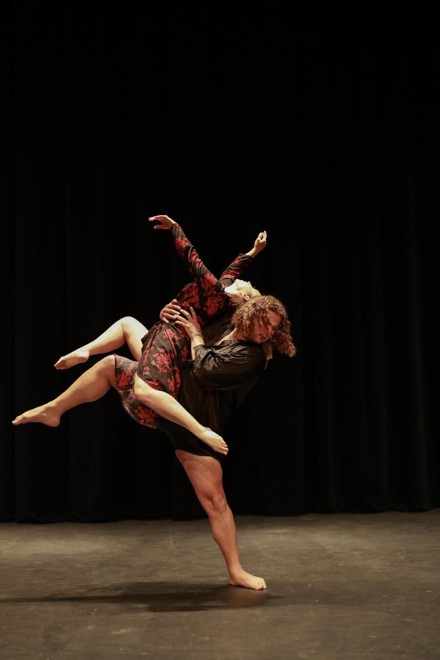 Two dancers mid lift on a dark stage, one carrying the other horizontally across the body.