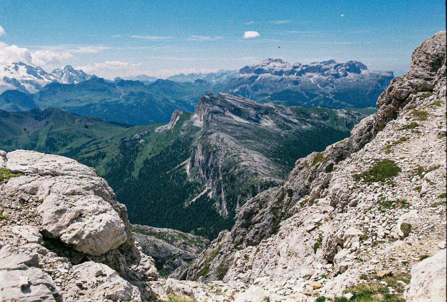 A steep rocky descent framing a long green valley, with a line of blue peaks far beyond.