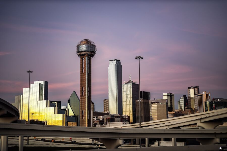 The Dallas skyline at dusk behind a highway overpass, with Reunion Tower lit against a violet sky.