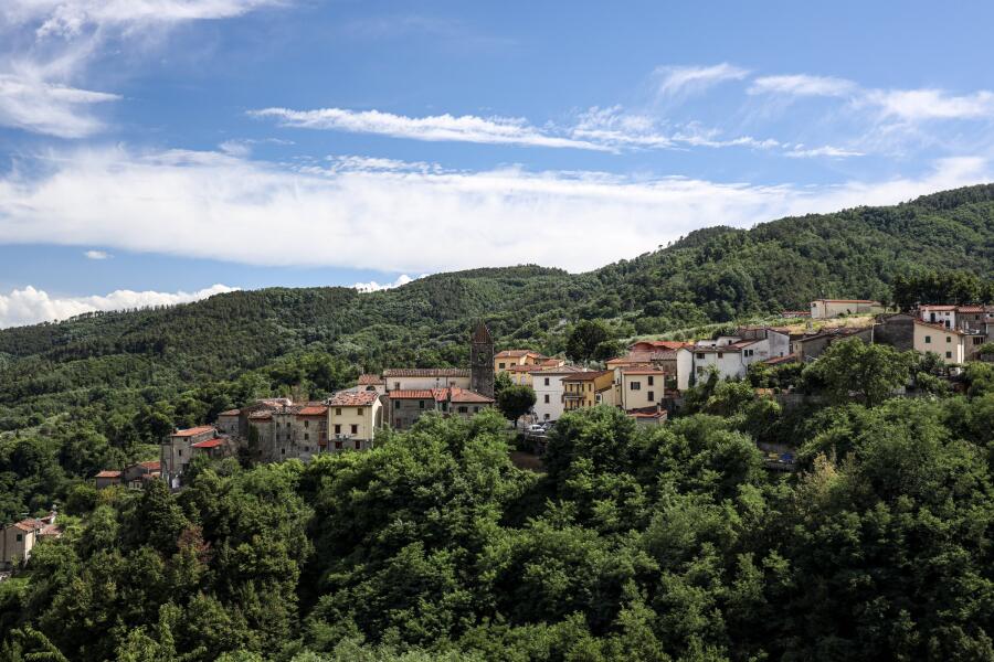 A small hill town of stone houses seen across a wooded valley under a wide sky of broken cloud.