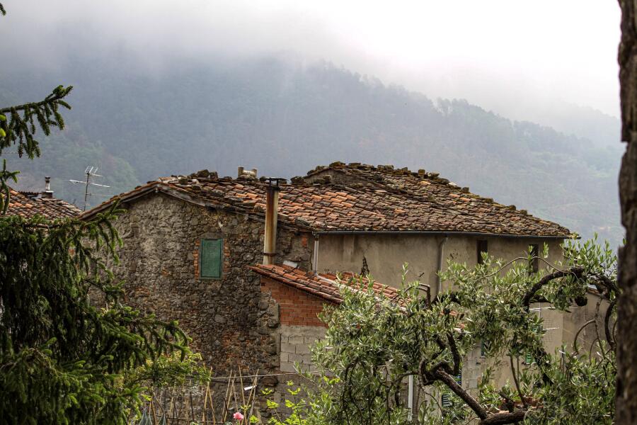 An old stone house with a heavy tiled roof, backed by a mountain slope disappearing into mist.