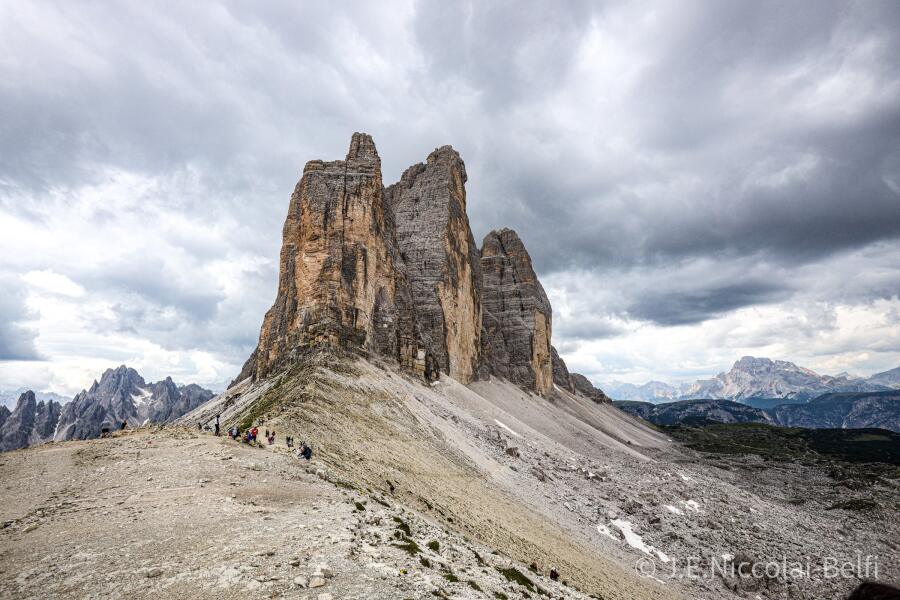 Three tall rock towers rising side by side above a pale scree ridge under heavy cloud.