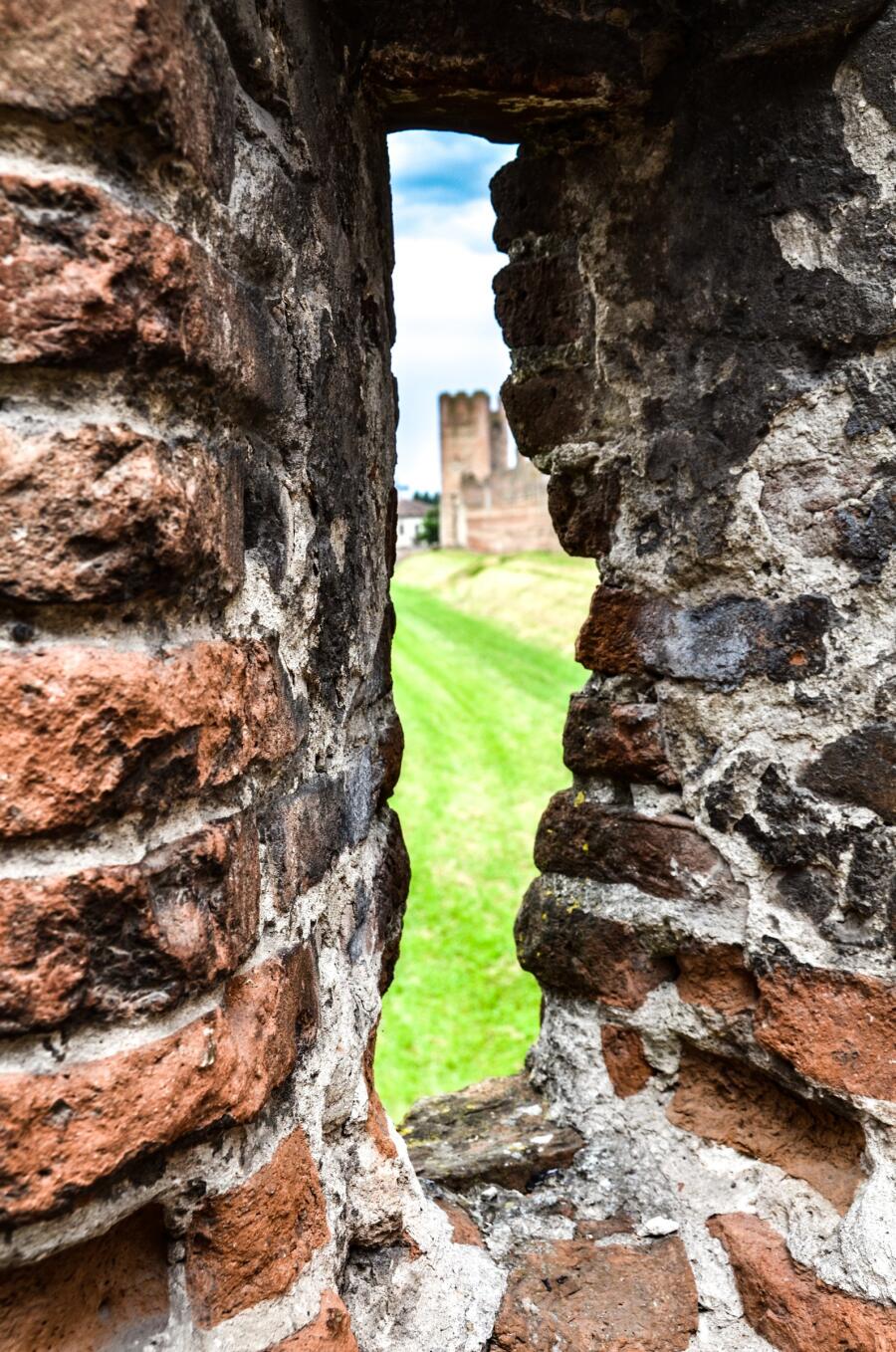 A tower and bright green field framed through a narrow slit window in a thick stone wall.