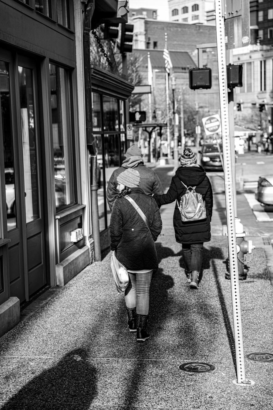 Black and white street photograph of two people walking away down a narrow city sidewalk past shopfronts.