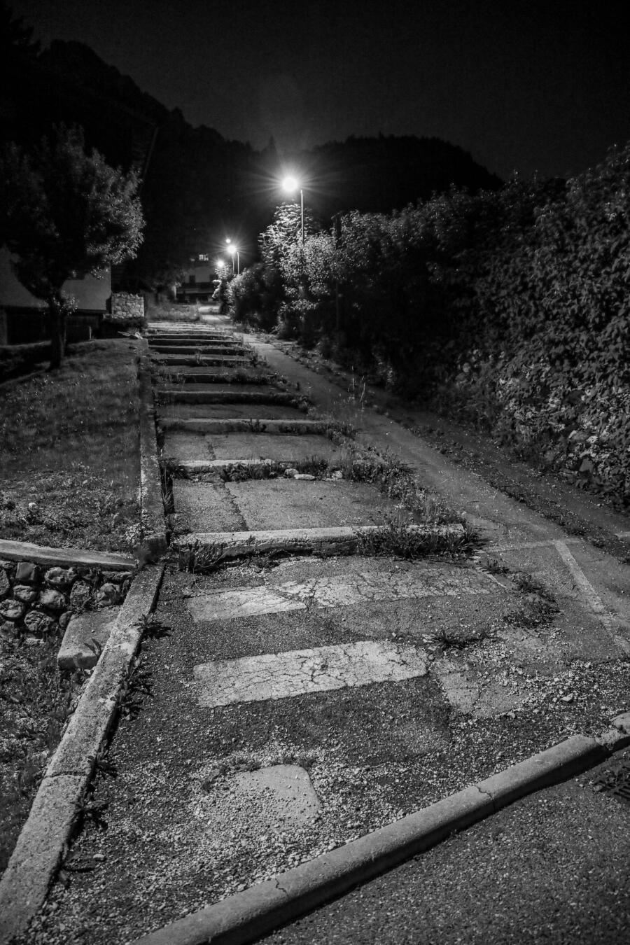 Black and white photograph of a rough road at night, lit by a single street lamp between dark hedges.